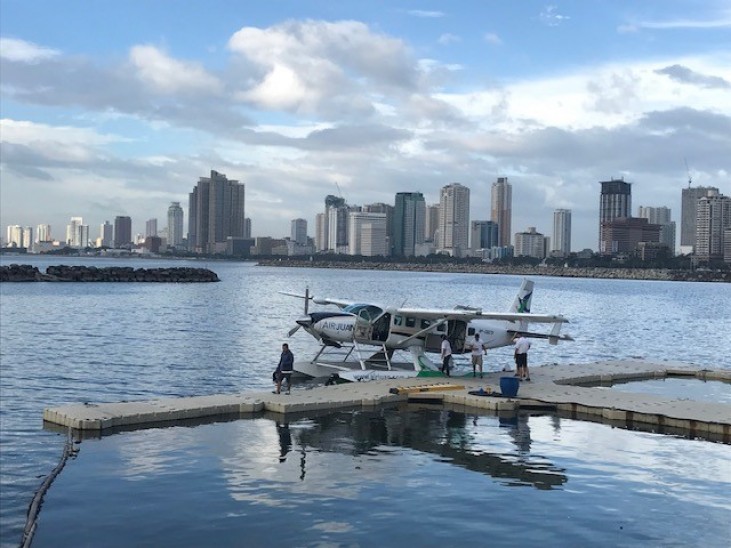 A seaplane is moored at a floating pier in a calm bay, with several people nearby. Tall city buildings and a cloudy blue sky form the background across the water.