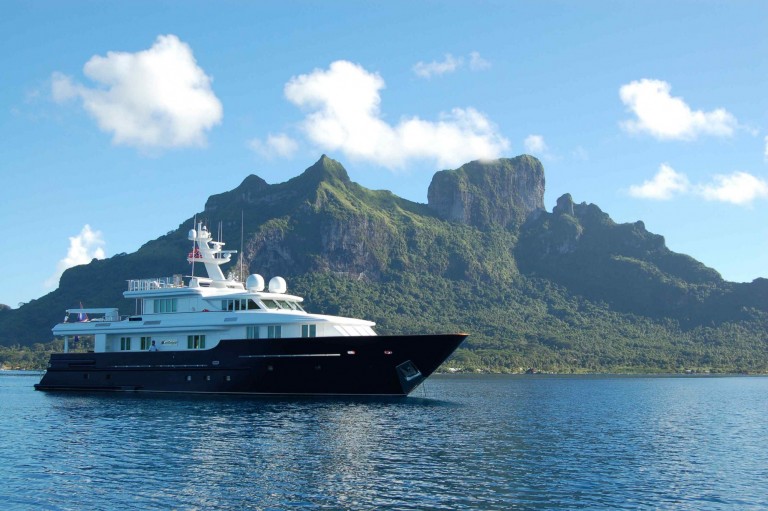 A large luxury yacht floats on calm blue water with a lush, green mountain and a bright blue sky with scattered clouds in the background.