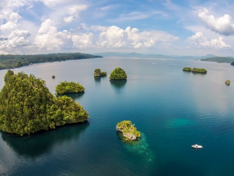 Aerial view of a calm blue sea with several small, lush green islands scattered throughout. A small white boat floats near one island, and the sky is filled with fluffy white clouds.