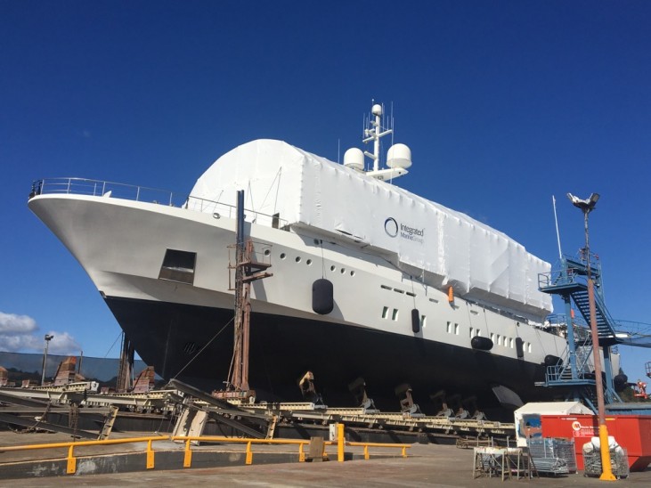 A large yacht with a white covered upper deck sits on supports in a shipyard under a clear blue sky. Yellow barriers and industrial equipment are visible around the vessel.