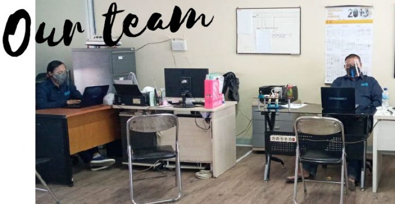 Two people wearing masks work at desks in an office with computers and office supplies. The words Our team appear in large text at the top left. Empty chairs are placed between the desks.