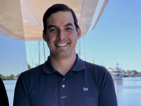 A man with dark hair, wearing a navy polo shirt with YM on it, smiles whilst standing in front of a marina with boats and blue sky in the background.
