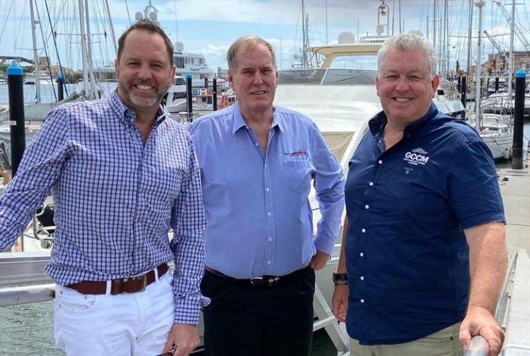 Three men stand smiling at a marina in front of boats and yachts. All are wearing collared shirts; the man on the left wears white trousers, the others wear dark trousers. The background features water, masts, and a partly cloudy sky.