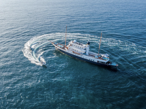 A large yacht is anchored in the sea, whilst a small speedboat circles around it, leaving a visible trail in the water. The scene is viewed from above.