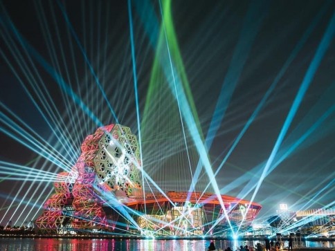 A modern, geometric building by the water is illuminated with colourful, intersecting laser lights in the night sky, creating a vibrant and dynamic scene reflected on the water’s surface.