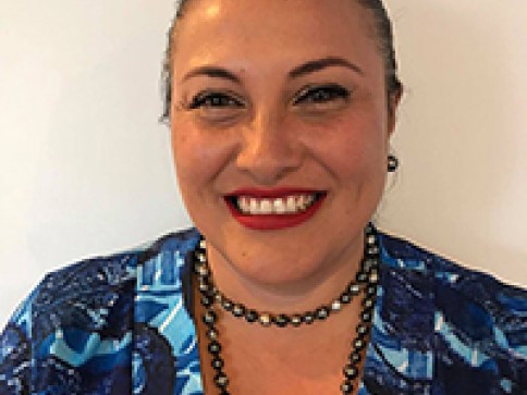 A woman with dark hair pulled back, wearing a bright blue patterned top, red lipstick, and a black beaded necklace, smiles at the camera against a plain light background.