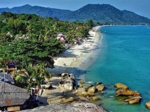 A tropical beach with turquoise water, large rocks, white sand, and palm trees. Small huts and lush green hills are in the background under a sunny, clear blue sky. People are relaxing on the beach and swimming in the sea.