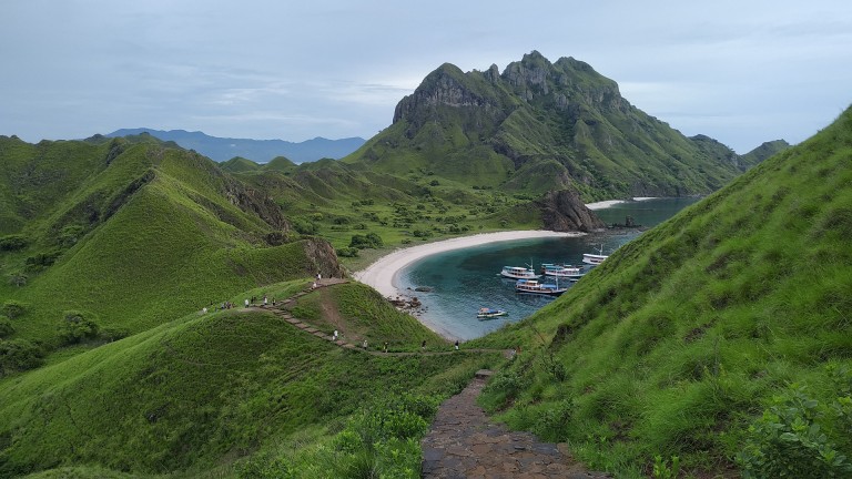 A lush green hill overlooks a curved sandy beach and turquoise bay, with several boats anchored near the shore. Tall, rugged mountains rise in the background under a cloudy sky.