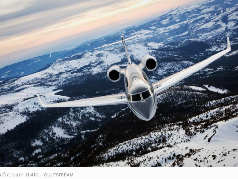 A sleek Gulfstream G500 jet flies over snow-covered mountains and forests under a cloudy sky, showcasing its modern design against a dramatic winter landscape.