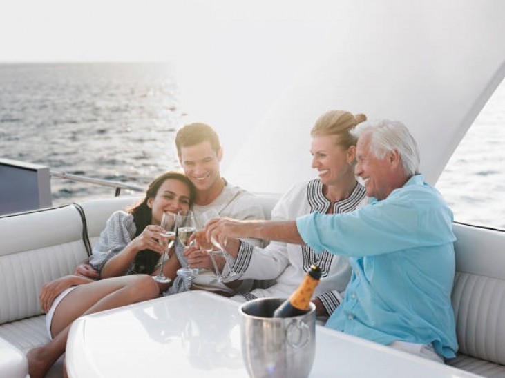 Four people sit together on a yacht, smiling and enjoying glasses of champagne. The sea is visible in the background, and a bottle of champagne rests in an ice bucket on the table in front of them.