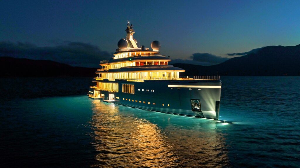 A large, luxurious yacht brightly illuminated by lights cruises on calm waters at dusk, with dark mountains and a cloudy sky in the background.