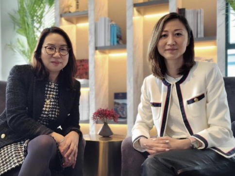 Two women are sitting indoors on chairs next to a small table with a vase of red flowers. Shelves with books and plants are in the background. Both women are looking at the camera and smiling slightly.