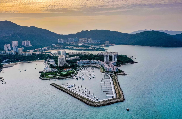 Aerial view of a modern marina with yachts moored, surrounded by residential buildings and green hills, bordered by calm blue water and backed by a dramatic sky at sunset.