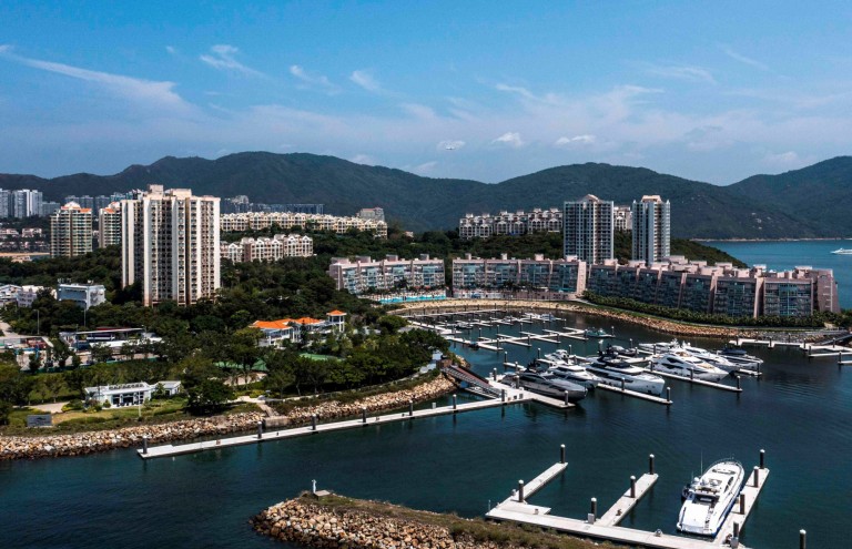 A marina with several yachts and boats moored near modern blocks of flats, surrounded by greenery and mountains under a clear blue sky.