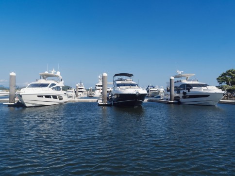 Three luxury yachts are moored at a marina on a clear, sunny day. The water is calm, and several other boats are visible in the background, with blue sky overhead.