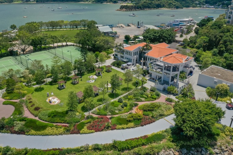 Aerial view of a park with green lawns, walking paths, gazebos, tennis courts, and a white building with an orange roof, located near a waterfront with boats and trees.