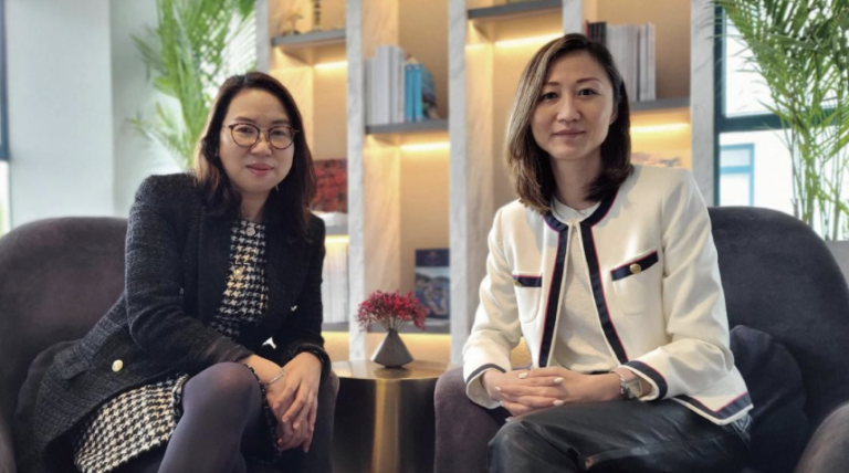 Two women sit in armchairs beside a small round table with a plant, in a modern office with bookshelves and greenery in the background. Both are smiling and dressed in business attire.