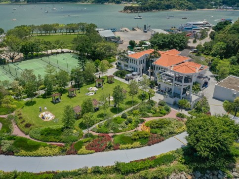 Aerial view of a lush green park with walking paths, gazebos, and gardens next to a white building with an orange roof, tennis courts, and a waterfront with boats and trees in the background.