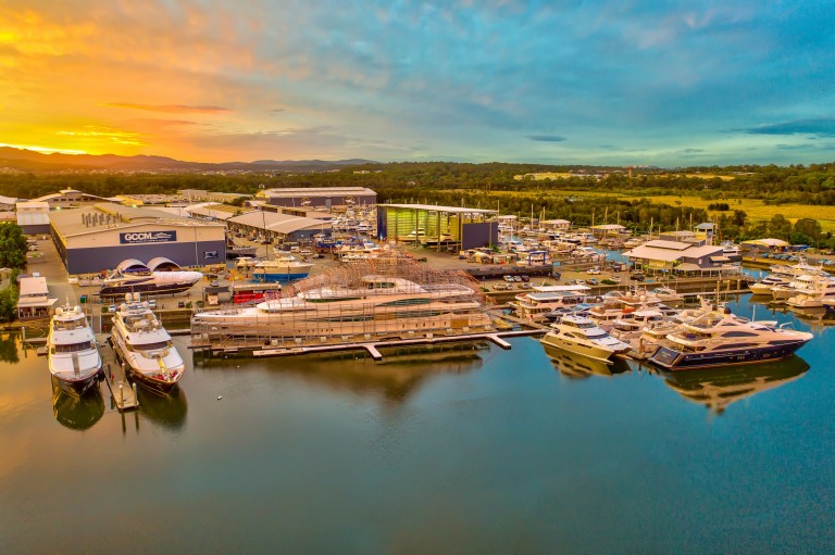 Aerial view of a marina at sunset with yachts and boats moored, boat sheds, and green surroundings. The water reflects the vibrant sky with orange and blue hues.