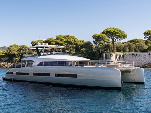A modern, luxury catamaran yacht is anchored on calm blue water near a lush, tree-lined shore under a clear sky.