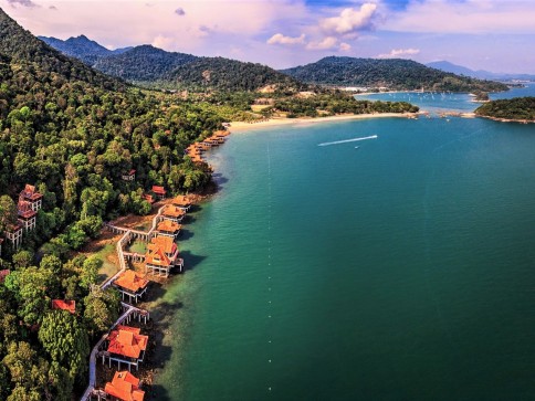 Aerial view of a tropical coastline with lush green hills, turquoise water, villas with red roofs along the shore, and boats creating white trails on the water under a partly cloudy sky.