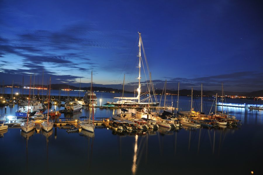 A marina at dusk with several sailboats and yachts moored in calm water, reflecting lights from the boats and nearby buildings under a deep blue sky.