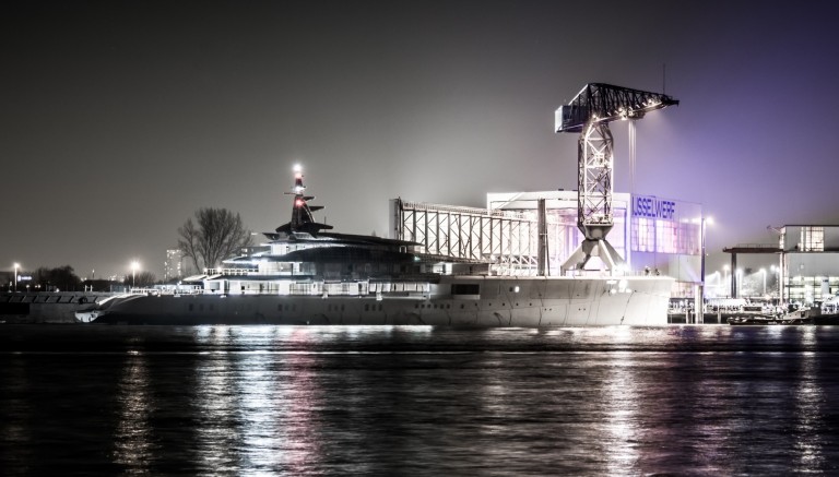 A large, modern yacht is moored at a shipyard at night, with industrial cranes and illuminated buildings reflected in the calm water.