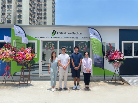 Four people stand in front of a Lodestone Yachts building with flower arrangements on each side. The background includes a city building and clear blue sky on a sunny day.