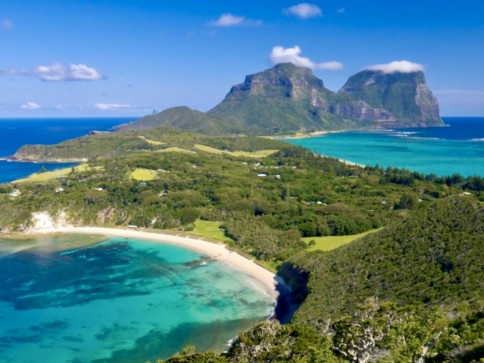 A scenic view of a turquoise bay, lush green hills, and a sandy beach, with two steep, rocky mountains rising in the background under a clear blue sky.