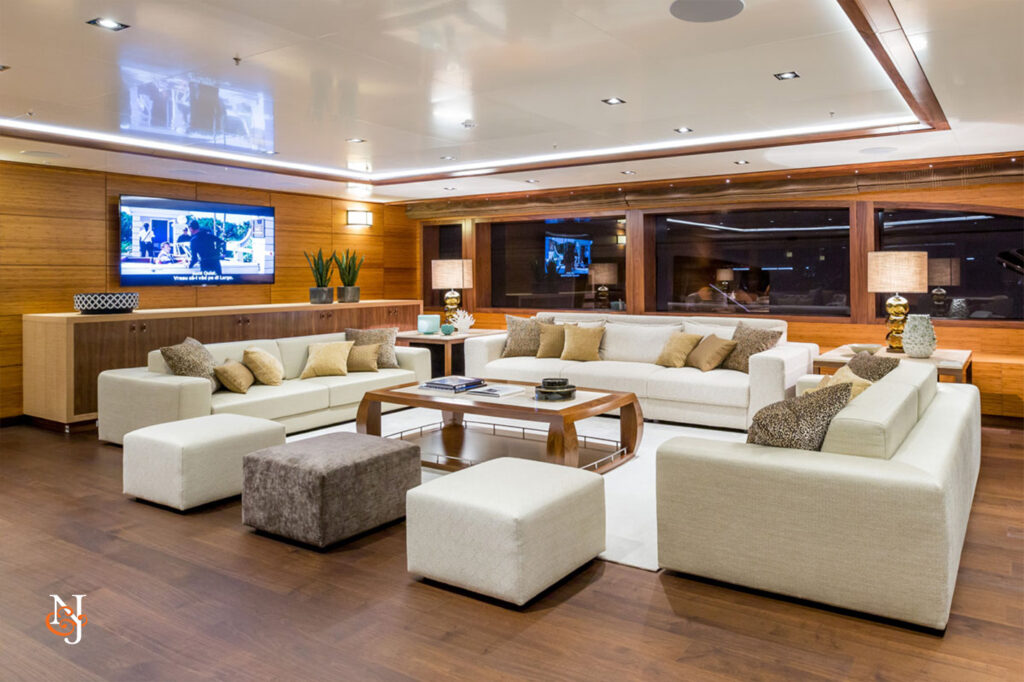 Modern yacht lounge with cream sofas, ottomans, and wooden accents. Large windows line one wall, offering a view outside. TVs are mounted, and decorative plants and lamps add a cosy, upmarket atmosphere.
