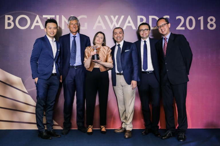 Six people dressed in formal attire stand together smiling at the Boating Awards 2018 event; one woman in the centre holds a trophy, with a purple backdrop behind them displaying the event name.