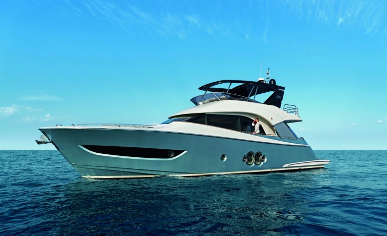A modern luxury yacht with sleek design floats on calm blue sea under a clear sky. The yacht has large windows, a spacious upper deck, and a person is visible at the helm.