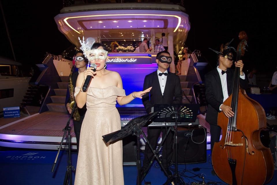 A woman in an elegant dress and white mask sings into a microphone beside musicians in masks and formal wear, performing on a stage in front of a brightly lit yacht at night.