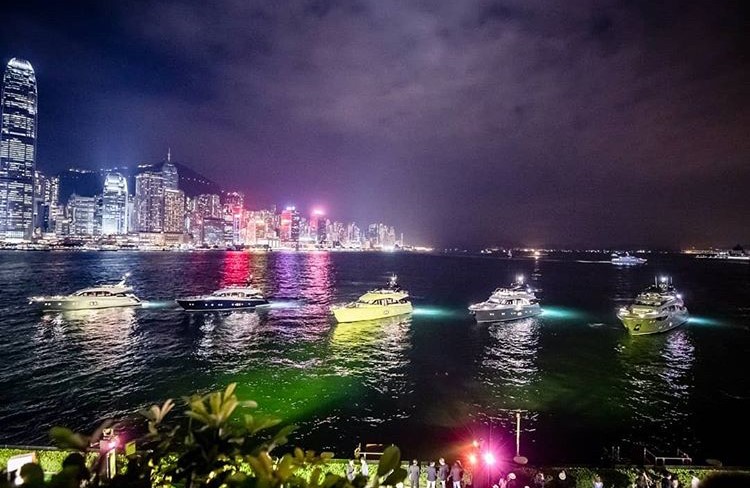 A nighttime cityscape with illuminated skyscrapers along a waterfront. Several yachts with glowing underwater lights float on the water, reflecting city lights. People watch from a garden in the foreground.
