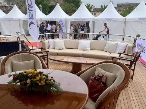 A yacht deck with a round table, floral centrepiece, chairs, and a beige sofa with white cushions. People and white marquees are visible in the background at an outdoor event. A red handbag sits on one chair.