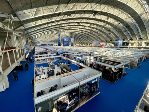 A large indoor trade fair with blue carpet, many exhibition stands, and a curved, vaulted ceiling. People walk between stands displaying products and banners under bright lighting.