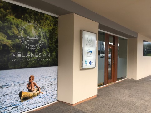 A shopfront with large signs for Melanesian Luxury Yacht Services, including a photo of a child paddling a canoe on water with green foliage in the background. Glass doors and a sign with multiple business logos are visible.