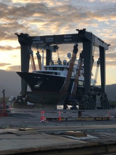 A large boat is being lifted out of the water by a massive mobile boat hoist at a shipyard, with the sun rising or setting in the background and mountains visible in the distance.