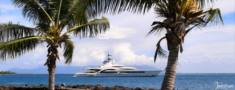 A large white yacht floats on blue water, framed by two palm trees in the foreground. Dark rocks and tropical greenery are visible, with a cloudy sky and distant islands in the background.