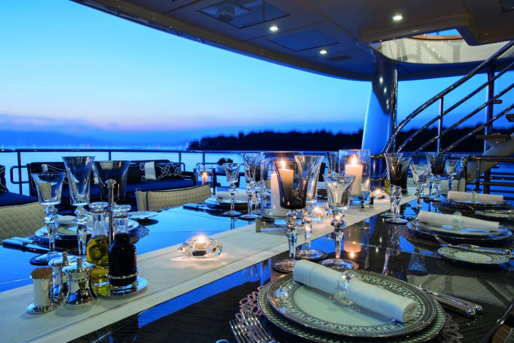 Elegant outdoor dining table set with crystal glasses, cutlery, and candles on a yacht at sunset, overlooking calm water and a distant shoreline, with a modern spiral staircase visible on the right.