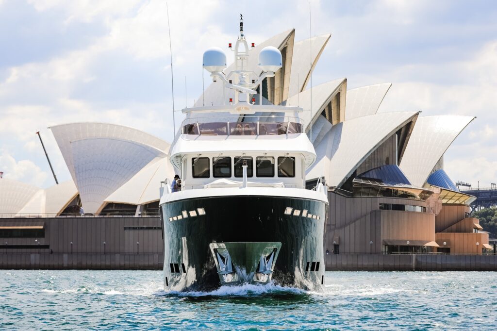 A large yacht sails on the water in front of the Sydney Opera House on a partly cloudy day. The iconic white sails of the Opera House are visible in the background.