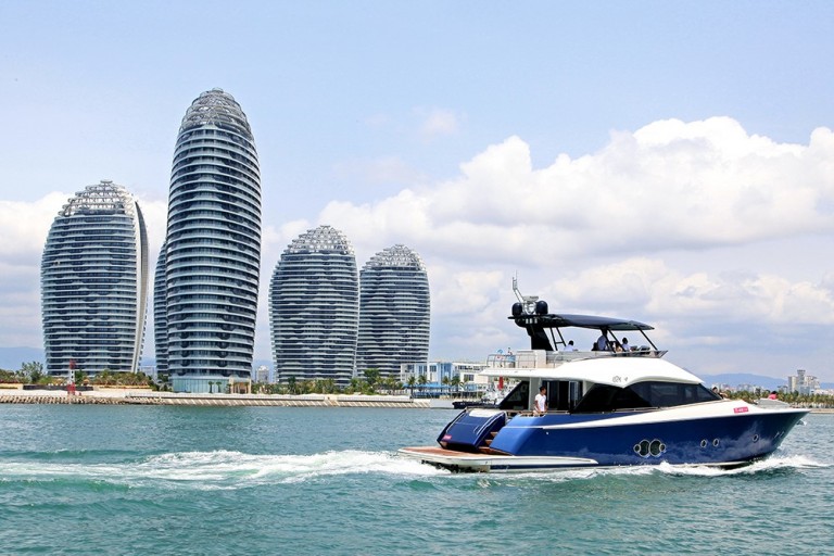 A blue and white yacht sails on the water with several tall, modern, oval-shaped buildings in the background under a partly cloudy sky.