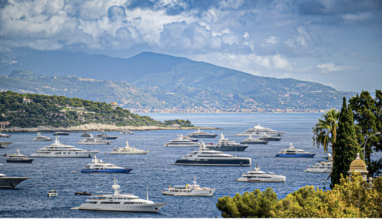 Numerous luxury yachts anchored in a bay with green hills and mountains in the background, under a cloudy sky. Trees and greenery frame the foreground.