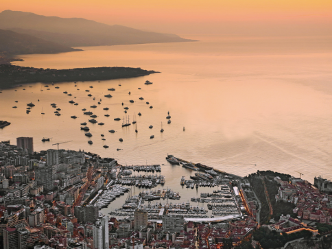 Aerial view of a coastal city at sunset, with numerous boats anchored in a calm bay and urban buildings densely packed along the waterfront, bordered by mountains in the background.