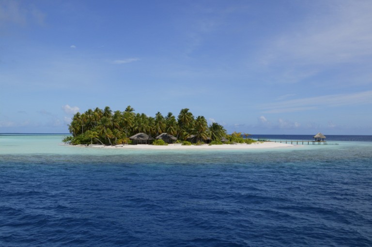 A small tropical island with palm trees, a few thatched-roof huts, and a wooden pier sits surrounded by turquoise shallow water and deep blue sea under a bright, partly cloudy sky.