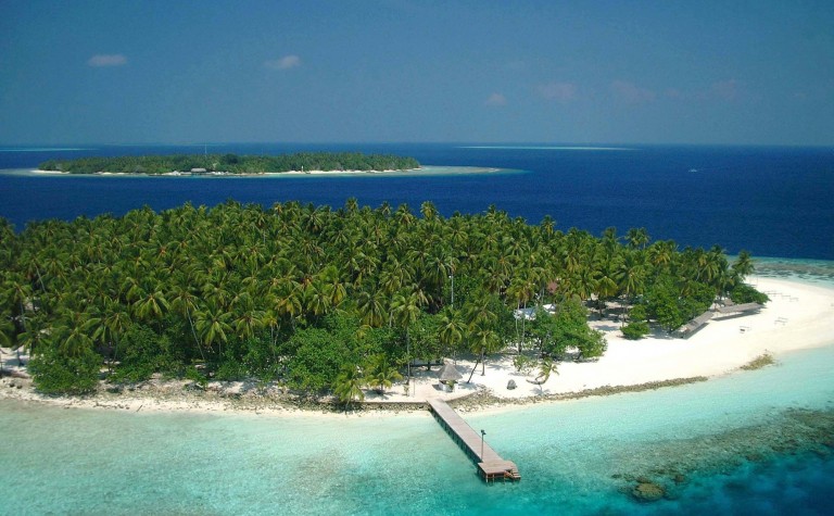 A tropical island with lush green palm trees, white sandy beaches, and a wooden jetty extending into clear turquoise water, with another island visible in the distance across the blue sea.