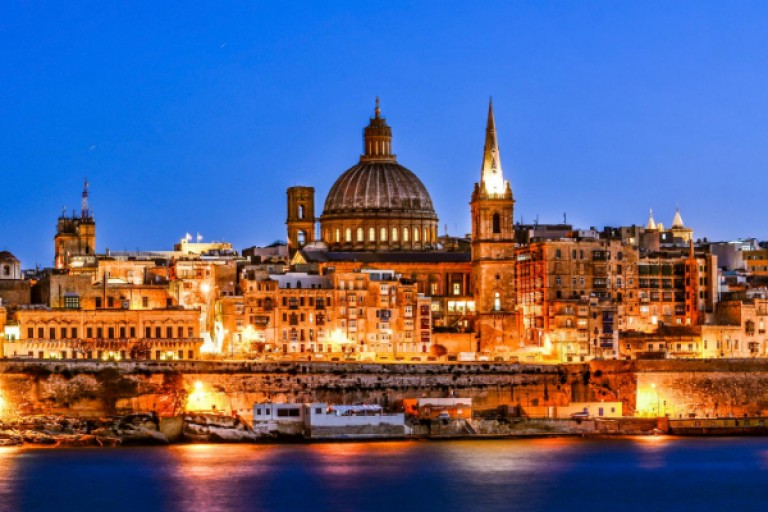 A brightly lit cityscape at dusk shows historic buildings and a large domed church, with their reflections shimmering on the calm waterfront. The sky is deep blue, creating a striking contrast with the warm city lights.