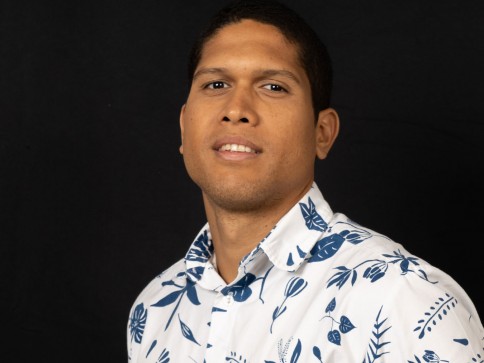 A man with short dark hair wearing a white shirt with blue floral patterns poses in front of a plain black background, looking slightly to the side and smiling softly.