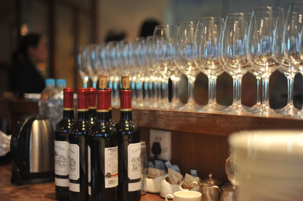 Several bottles of red wine are lined up on a counter next to a row of empty wine glasses, with a kettle, cups, and other items also visible. People are blurred in the background.