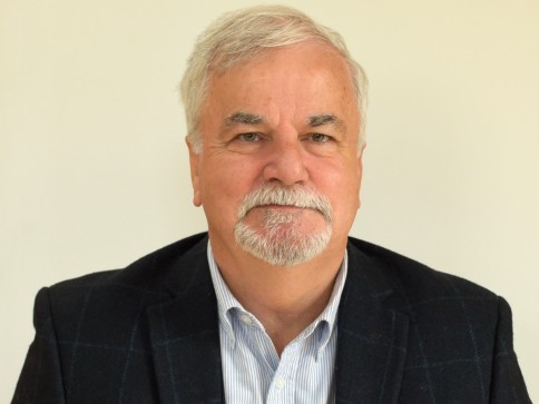 A man with short white hair, a moustache, and a trimmed beard is wearing a dark blazer and a light blue collared shirt, posing in front of a plain light background.